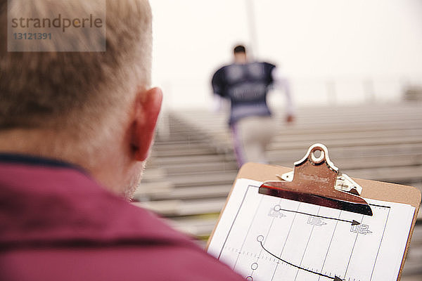 Rückansicht des Trainers  der die Zwischenablage hält  mit Blick auf den im Stadion trainierenden American-Football-Spieler