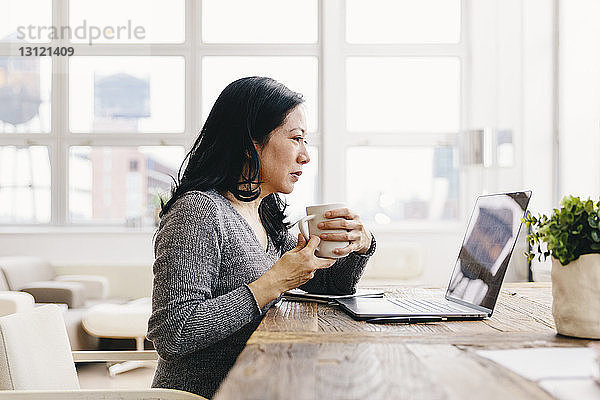 Geschäftsfrau hält Kaffeetasse in der Hand  während sie auf einen Laptop-Computer auf dem Schreibtisch im Büro schaut