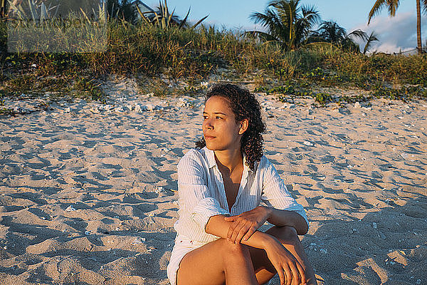 Frau schaut weg  während sie bei Sonnenuntergang am Strand auf Sand sitzt