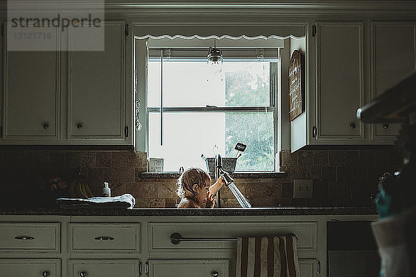 Verspieltes Mädchen  das mit dem Wasserhahn spielt  während es zu Hause am Fenster in der Küchenspüle sitzt