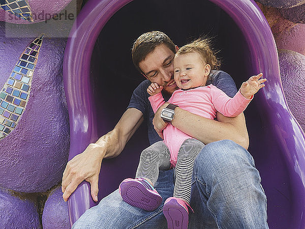 Vater mit süßer Tochter spielt auf dem Spielplatz auf einer Röhrenrutsche