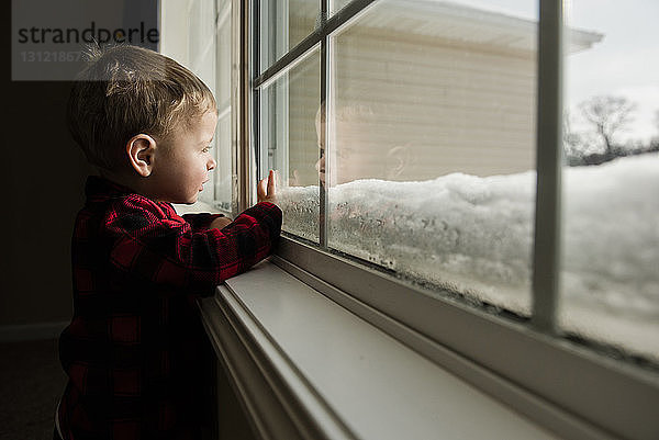 Seitenansicht eines Jungen  der durch ein Fenster schaut  während er im Winter zu Hause steht