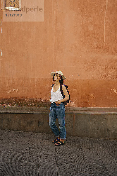 Porträt einer selbstbewussten Touristin in voller Länge mit Rucksack und Händen in den Taschen  die in der Stadt an der Wand steht