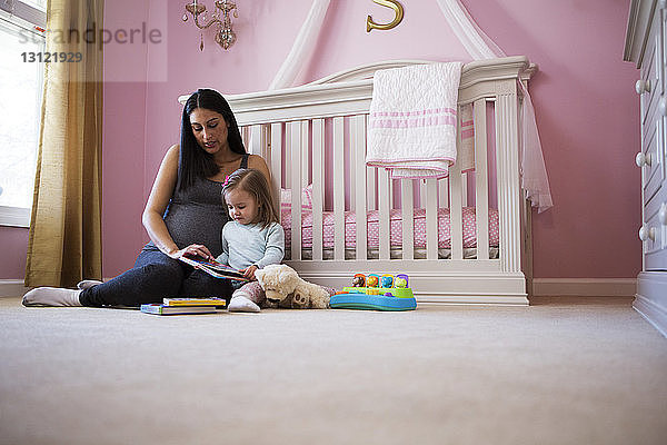 Schwangere Frau sitzt mit Tochter gegen Kinderbett zu Hause