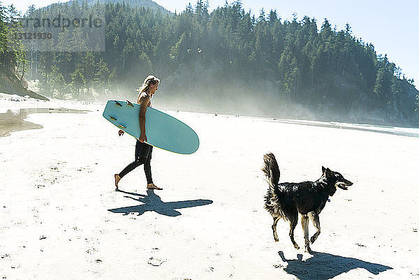 Frau hält Surfbrett mit Hund auf Sand am Strand spazieren