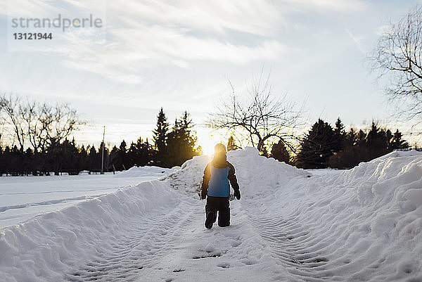 Rückansicht eines Jungen  der bei Sonnenuntergang auf einem schneebedeckten Feld gegen den Himmel läuft