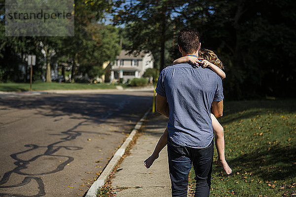 Rückansicht eines Vaters  der seine Tochter trägt  während er auf einem Fußweg geht