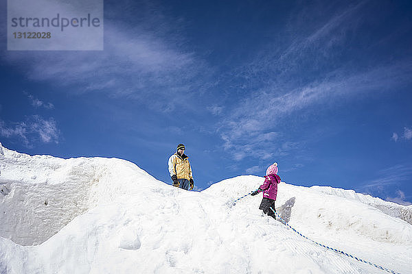 Niedrigwinkelaufnahme eines Vaters mit Tochter auf einem schneebedeckten Hügel vor blauem Himmel während eines sonnigen Tages