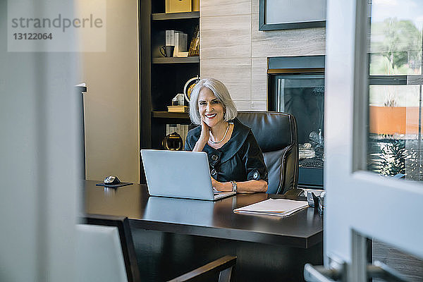 Porträt einer lächelnden Geschäftsfrau  die am Laptop-Computer am Schreibtisch im Büro arbeitet  durch die Tür gesehen