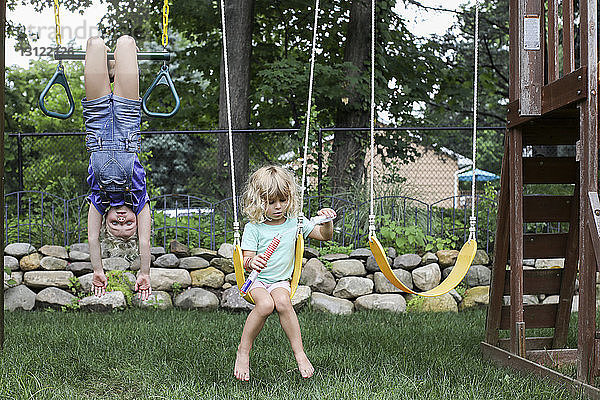 Mädchen hängt kopfüber  während die Schwester auf dem Spielplatz schaukelt