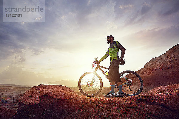 Wanderer in voller Länge mit Mountainbike auf Felsen stehend gegen den Wüstenhimmel bei Sonnenuntergang