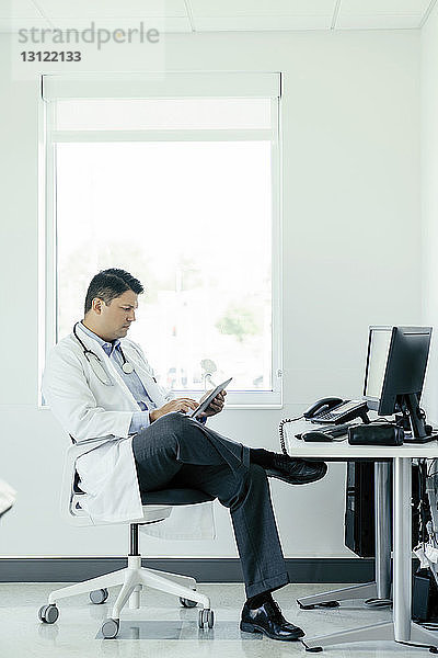 Arzt in voller Länge am Tablet-Computer  während er im Krankenhaus am Fenster sitzt