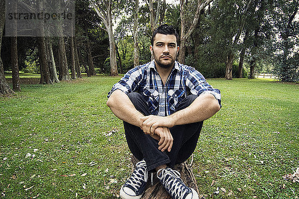 Porträt eines Mannes  der im Park auf Gras sitzend die Knie umarmt