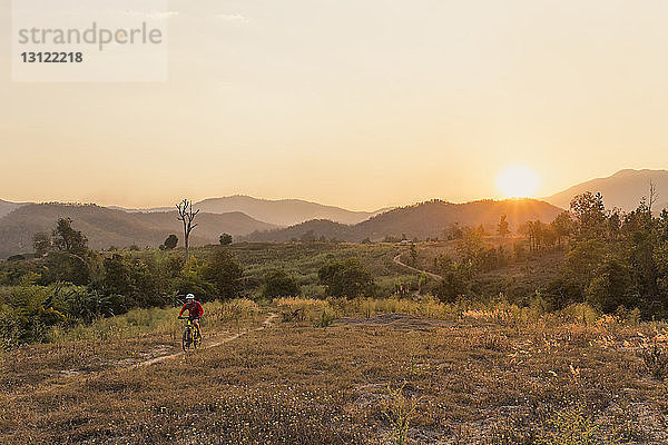 Hochwinkelansicht eines Mountainbike fahrenden Mannes auf dem Feld gegen den Himmel bei Sonnenuntergang