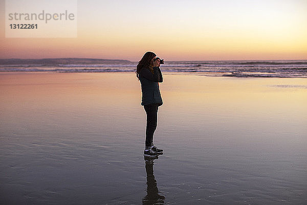 Seitenansicht einer Frau  die bei Sonnenuntergang am Strand steht und fotografiert