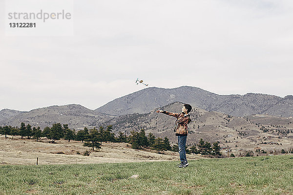 Seitenansicht eines Jungen  der ein Spielzeugflugzeug fliegt  während er auf einem Grasfeld gegen den Himmel steht