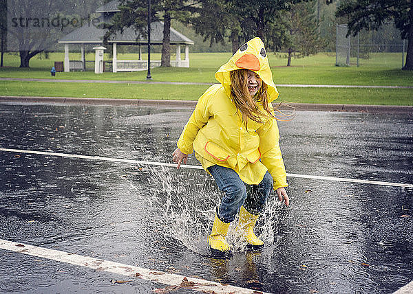 Verspieltes Mädchen in Regenmantel beim Springen in Pfütze bei Regen