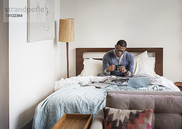 Mann hält Kaffeetasse  während er zu Hause im Bett Zeitung liest