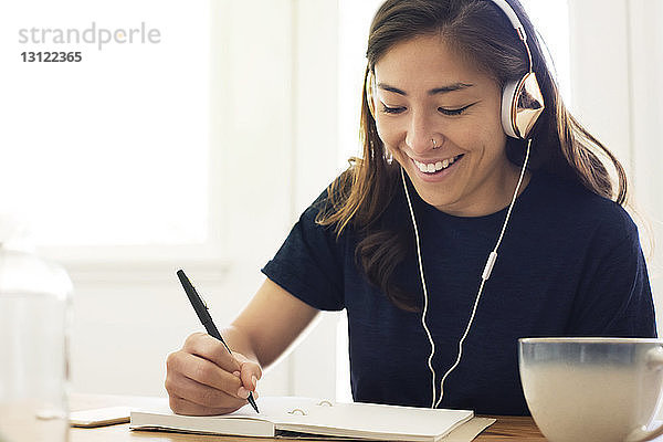 Fröhliche Frau hört Musik  während sie zu Hause in ein Buch schreibt