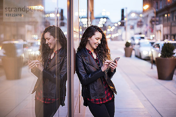 Lächelnde junge Frau  die ein Smartphone benutzt  während sie auf einem Fußweg an Gebäuden steht