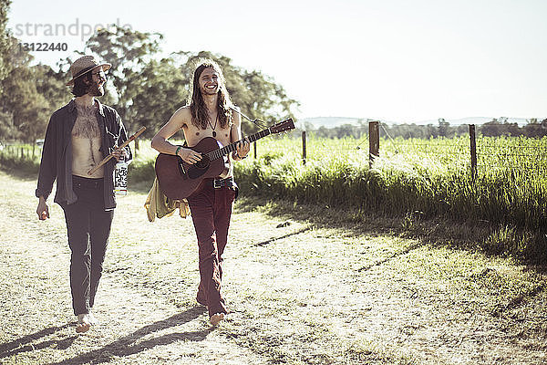 Glückliche Freunde spielen Musikinstrumente auf einem Feldweg durch ein Grasfeld gegen den Himmel
