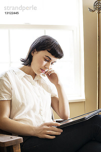 Frau liest digitales Tablet  während sie zu Hause am Fenster sitzt