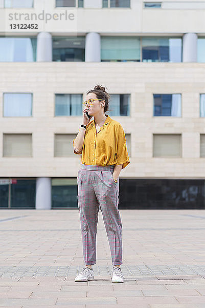 Selbstbewusste Geschäftsfrau spricht mit einem Smartphone  während sie auf dem Bürgersteig gegen ein Gebäude in der Stadt steht