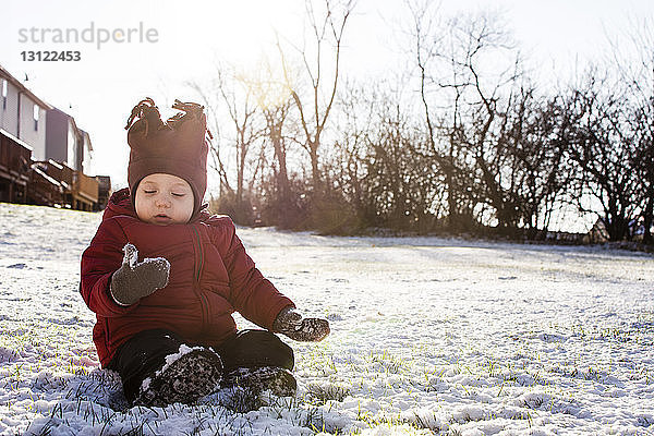 Junge in warmer Kleidung sitzt auf schneebedecktem Feld im Hinterhof