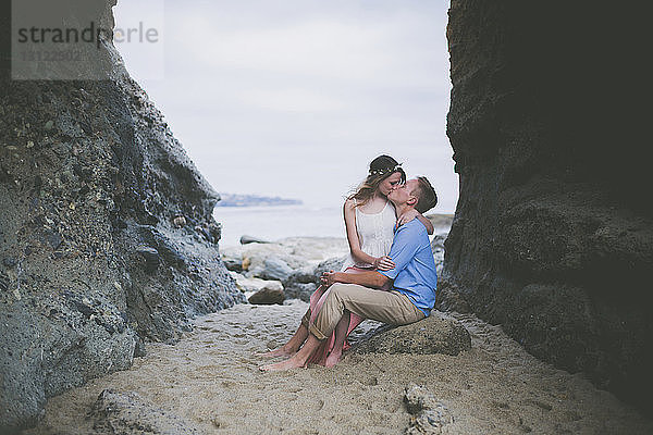 Romantisches Paar küsst sich am Strand auf einem Felsen sitzend