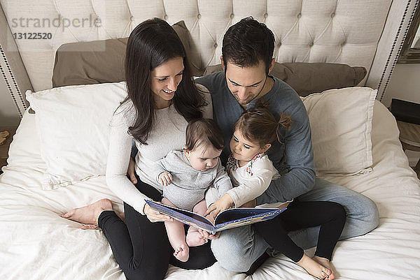 Hochwinkelansicht von Eltern mit Kindern  die auf dem Bett sitzend ein Bilderbuch betrachten