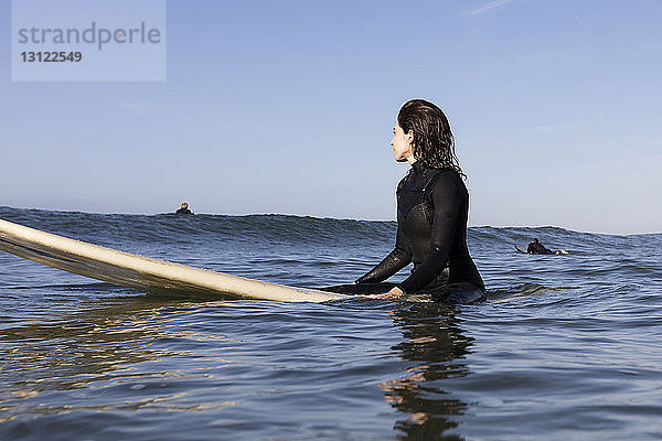 Nachdenkliche Surferin sitzt auf einem Surfbrett im Meer vor klarem Himmel
