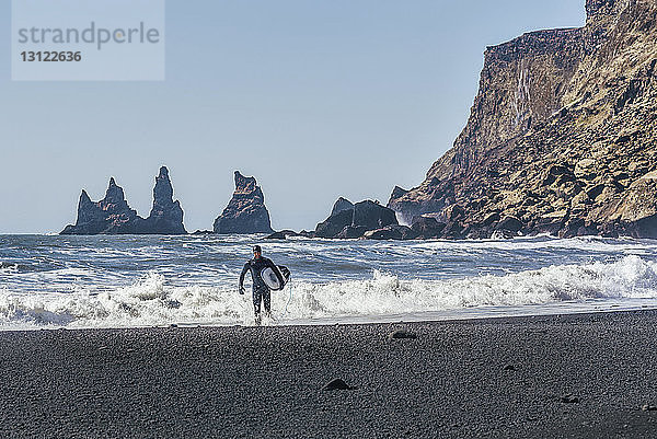 Mann mit Surfbrett am schwarzen Strand bei klarem Himmel am sonnigen Tag