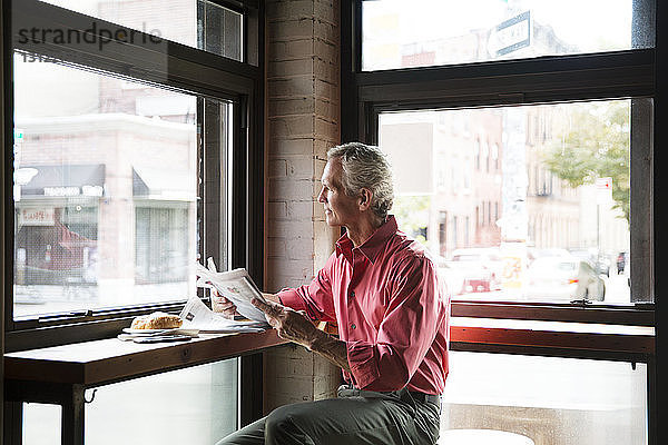Reifer Mann liest Zeitung  während er im Café ein Croissant isst