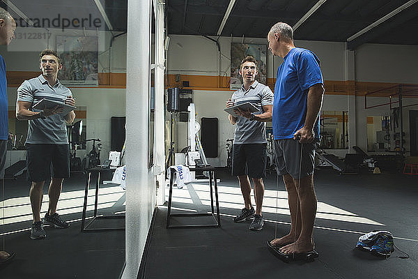 Trainer mit Tablet-Computer im Gespräch mit einem Mann  der im Fitnessstudio Dehnübungen macht