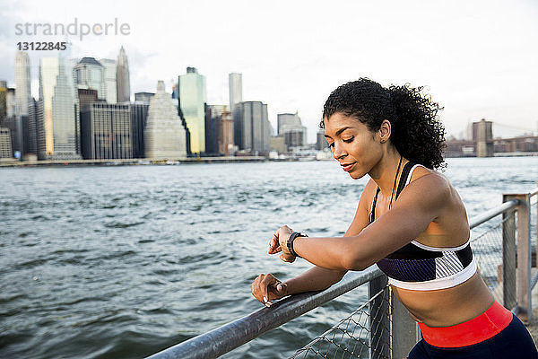 Weibliche Sportlerin prüft intelligente Uhr  während sie in der Stadt am Fluss gegen den Himmel steht