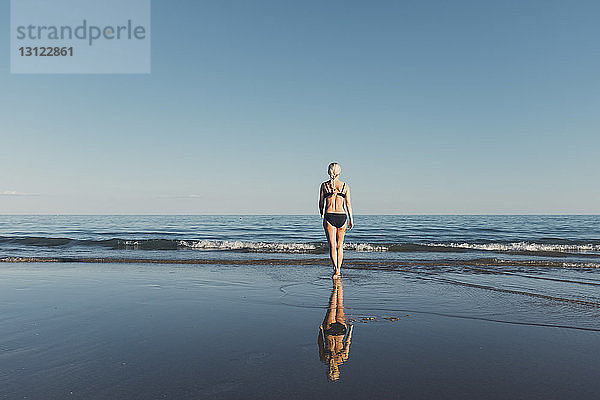 Rückansicht einer Frau im Bikini beim Spaziergang am Strand in Richtung Meer bei klarem Himmel