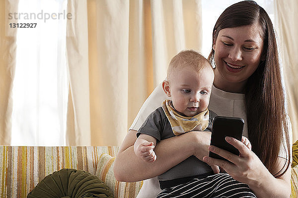 Glückliche Mutter zeigt dem Baby ihr Handy  während sie zu Hause auf dem Sofa sitzt