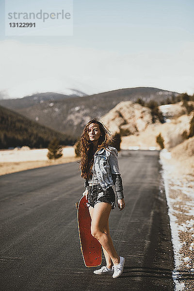 Junge Frau in voller Länge mit Skateboard  die an einem sonnigen Tag auf der Straße steht
