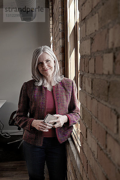 Porträt einer selbstbewussten  reifen Geschäftsfrau  die im Kreativbüro ein Smartphone am Fenster hält