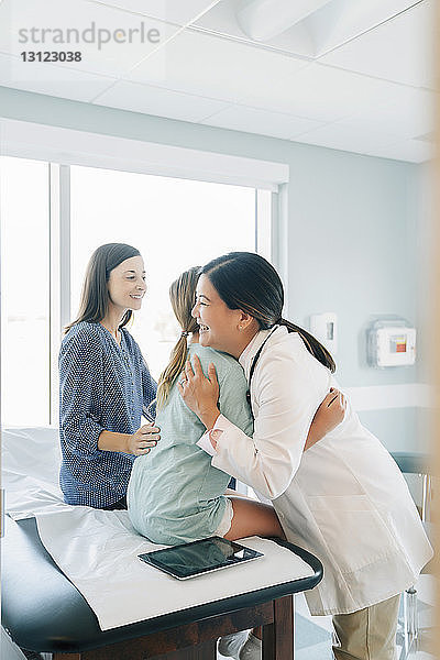 Fröhliche Kinderärztin umarmt Mädchen  das von der Mutter auf dem Untersuchungstisch im Krankenhaus sitzt
