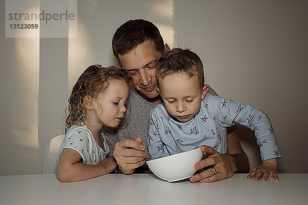 Vater füttert Kinder  während er zu Hause am Tisch an der Wand sitzt
