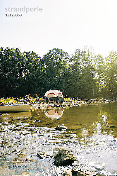 Mitteldistanzansicht von Freunden  die sich entspannen  während sie auf einem Campingplatz am See gegen den Himmel sitzen