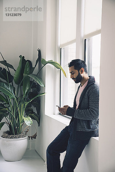 Geschäftsmann benutzt Smartphone  während er sich im Büro auf die Fensterbank lehnt