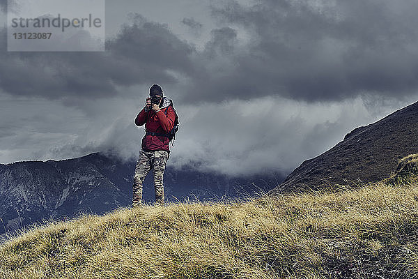 Männlicher Wanderer fotografiert im Stehen auf Berg vor bewölktem Himmel