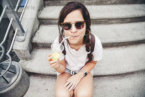 Hochwinkelporträt einer Frau mit Sonnenbrille beim Trinken von Fruchtsaft auf Stufen