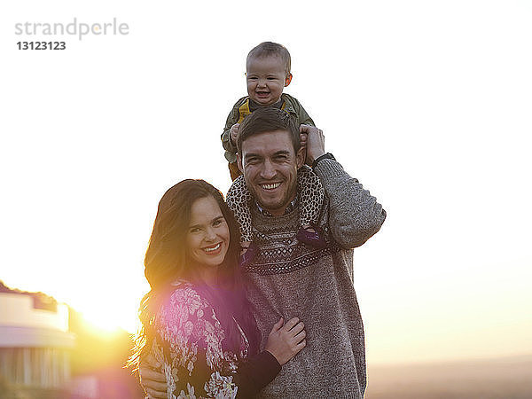 Porträt glücklicher Eltern mit niedlicher Tochter  die bei Sonnenuntergang am klaren Himmel stehen
