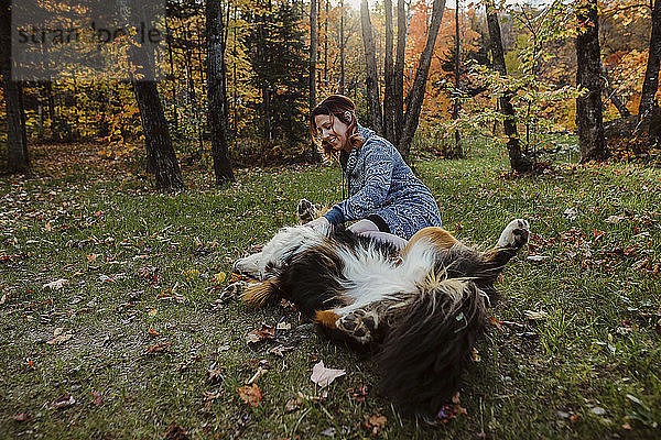 Fröhliche Frau spielt mit Hund  der im Herbst auf einem Grasfeld im Wald liegt