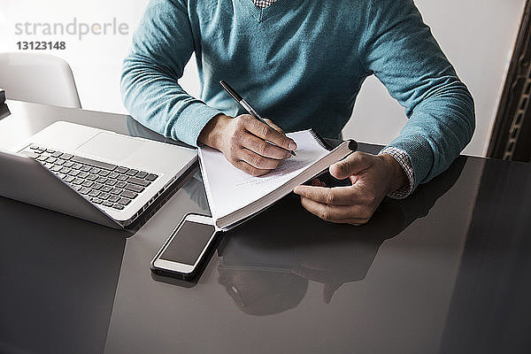 Mittelsektion eines Mannes  der mit einem Laptop an einem Tisch im Büro auf ein Buch schreibt