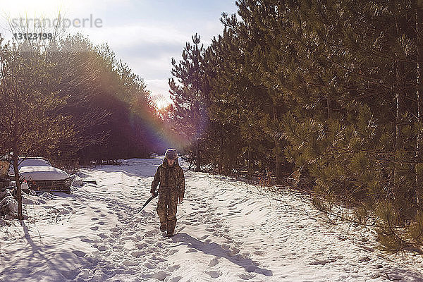 Junge in voller Länge mit Gewehr in der Hand auf schneebedecktem Feld inmitten von Bäumen