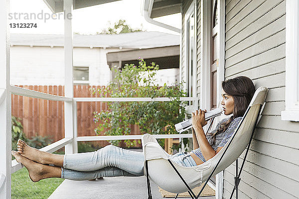 Seitenansicht einer Frau  die eine Zeitung in der Hand hält  während sie auf einem Stuhl auf der Veranda sitzt